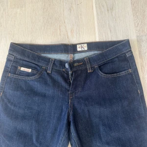  Lågmidjade Mörkblå bootcut jeans från Calvin Klein - Snygga mörkblå jeans från Calvin Klein med klassisk femficksdesign och bootcut-ben. Jeansen har kontrastsömmar och logga på fickan samt i midjan. Tillverkade i slitstarkt denimtyg som ger en tidlös look. Mijdemått 41cm rakt över innebensmått längd :88 cm