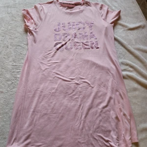Rosa sovklänning Juicy Couture - Söt rosa t-shirtklänning från Juicy Couture med glittrig text 'JUICY DRAMA QUEEN' framtill. Klänningen har korta ärmar och är gjord i mjuk bomull, perfekt för en chill och trendig look.