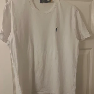 Vit t-shirt från Polo Ralph Lauren - Vit t-shirt från Polo Ralph Lauren med klassisk rund hals och korta ärmar. Diskret Polo-logga broderad på bröstet. Tillverkad i mjuk bomull som känns skön mot huden. Enkel och stilren design som funkar till allt.