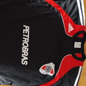 River Plate Adidas fotbollströja svart/röd - River Plate fotbollströja från Adidas i svart med röda och vita detaljer. Tryck med Petrobras på framsidan och klubbmärke på bröstet. RIVER-tryck på ryggen. Klassisk sportig design i lätt och ventilerande material.