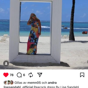 Färgglad långklänning från Lise Sandahl - Maxiklänning från Lise Sandahl med tropiskt blommönster i blått, rosa, gult och grönt. Ärmlös modell med rund halsringning, halterneck och markerad midja. Perfekt för sommarens alla tillfällen när du vill sticka ut med färg och mönster. Onesize.