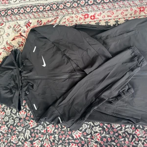 Nike Miller Windbreaker - Svart vindjacka från Nike med huva och hel dragkedja framtill. Jackan har reflekterande detaljer och elastiska muddar vid ärmsluten. Perfekt för blåsiga dagar och sportiga outfits. Swoosh-logga på bröstet ger en clean och sportig look.