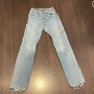 Levi's 501 ljusblå slitna jeans - Ljusblå Levi's 501 jeans med klassisk knappgylf, raka ben och slitna detaljer på både fram- och baksida. Råa, fransiga benslut och flera hål ger en riktigt vintage vibe. Perfekta för dig som gillar en avslappnad och edgy stil. Fina slitningar på låret samt bak vid rumpan. jätte fina men kommer inte till användning! 💓
