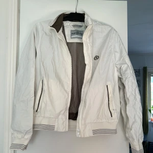 Vit vindjacka från Henri Lloyd - Snygg vit vindjacka från Henri Lloyd med brunt HL-logga broderat på bröstet. Jackan har dragkedja, två fickor med bruna detaljer och ribbade muddar med ränder. Perfekt för vår och höst när du vill ha en clean och sportig look.