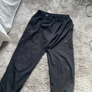 Svarta träningsbyxor från Nike - Svarta träningsbyxor från Nike med elastisk midja och diskret Nike-logga på benet. Byxorna har en rak passform och är tillverkade i ett lätt och smidigt syntetmaterial som passar perfekt för sport och träning.