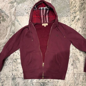 Vinröd ziphoodie från Burberry  - Säljer min vinröda hoodie från Burberry med klassiskt rutigt foder i huvan, guldfärgad dragkedja och broderad logga på bröstet. Perfekt för en snygg och avslappnad stil med ett exklusivt märke. Passar förmodligen också storlek XS!