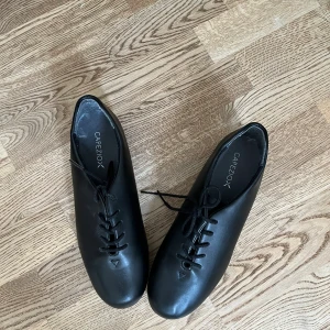 Svarta steppskor Capezio i skinn - Säljer ett par svarta steppskor från Capezio i äkta skinn. Skorna har snörning, rund tå och metallplattor under sulan för klassiskt stepp-ljud. Perfekta för danslektioner eller scenframträdanden. Stilren och tidlös design. Endast använt en gång!