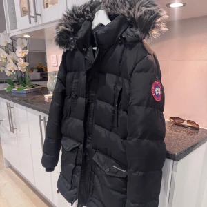 Svart dunjacka Canada Goose S - Säljer en svart dunjacka från Canada Goose i storlek S. Jackan har en fluffig pälskantad huva, flera praktiska fickor och klassisk patch på ärmen. Den är quiltad och har dragkedja framtill. Perfekt för kalla vinterdagar och riktigt snygg streetstil. Nästan som ny!