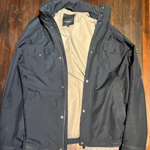 Svart vindjacka från Rockandblue - Säljer en svart Lex Tech vindjacka från Rockandblue. Jackan har två bröstfickor med knapp, dragkedja och tryckknappar framtill samt hög krage. Insidan är ljusbeige och materialet är slitstark polyester. Perfekt för dig som vill ha en stilren och funktionell jacka.