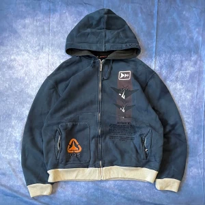 Avirex Vintage Hoodie – Rare Design - Classic Avirex full-zip hoodie with unique graphics and embroidery details. Features aircraft graphic print on the front, embroidered “1975” on the pocket, and branded zipper pull. Thick cotton material with ribbed cuffs and hem in contrasting beige.  📏 Measurements:  Length: 71 cm Chest (pit to pit): 62 cm Sleeve length: 81 cm  #Avirex #VintageHoodie #Streetwear #Y2KStyle #MilitaryFashion #RareFind #AviatorStyle #ArchiveFashion