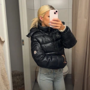 Svart Moncler pufferjacka med huva - Säljer en svart pufferjacka från Moncler med huva och dragkedja. Jackan är kortare i modellen och har Moncler-logga på ärmen samt coola detaljer som fickor med dragkedja. Materialet är glansigt och fyllningen är dun, perfekt för kalla dagar. Fråga om fler bilder om så behövs! 