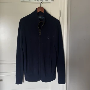 Mörkblå kofta från Polo Ralph Lauren - Klassisk mörkblå kofta från Polo Ralph Lauren med dragkedja framtill och broderad logga på bröstet. Tröjan är långärmad, har ribbade muddar och är tillverkad i mjuk bomull. Perfekt för lager-på-lager och snygg till både jeans och chinos.