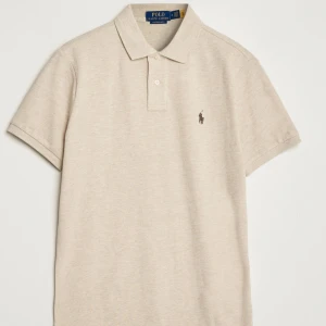 Beige pikétröja från Polo Ralph Lauren - Klassisk beige pikétröja från Polo Ralph Lauren med korta ärmar och broderad logga på bröstet. Tröjan har krage och två knappar framtill. Tillverkad i mjuk bomull för en skön och stilren look.