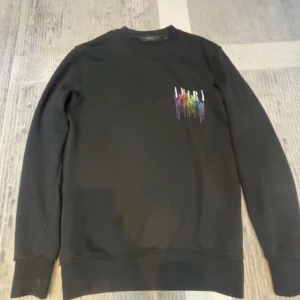 Svart Amiri sweatshirt med färgglatt tryck - Svart sweatshirt från Amiri med färgglatt, droppande logotryck på bröstet och ryggen. Tröjan har rund hals, långa ärmar och en relaxed fit. Perfekt för dig som vill sticka ut med en cool streetstyle-look.
