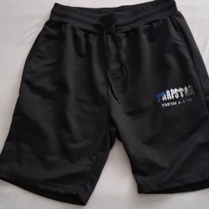 Svarta Trapstar shorts med tryck - Svarta shorts från Trapstar med tryck i blått, vitt och grått på ena benet. Mjuk bomullskänsla, elastisk midja med snörning och avslappnad passform. Perfekta för chill dagar eller streetwear-stil.