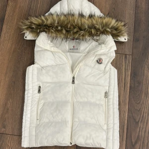 Vit dunväst från Moncler - Säljer en snygg vit dunväst från Moncler med quiltad design och hög krage. Västen har två fickor med dragkedja och Moncler-logga på bröstet. Materialet är polyester och dun, perfekt för kyliga dagar. Stängs med dragkedja framtill.
