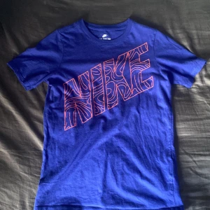 Blå Nike t-shirt med grafiskt tryck - Snygg blå t-shirt från Nike med stort grafiskt Nike-tryck i rött på bröstet. Klassisk rund hals och korta ärmar. Tillverkad i mjuk bomull som är skön att ha på sig hela dagen. Perfekt för dig som gillar sportig och avslappnad stil.