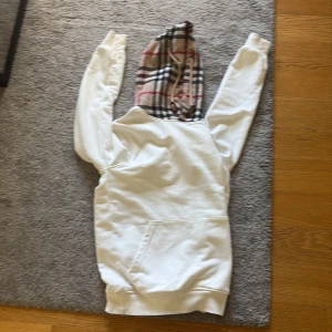 Vit hoodie från Burberry med rutig huva - Vit hoodie från Burberry med klassisk rutig huva i beige, svart och rött. Tröjan har lång ärm, känguruficka framtill och är tillverkad i mjuk bomull. Perfekt för dig som vill ha en stilren men ändå lyxig look.
