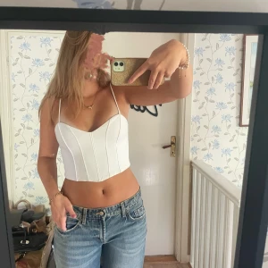 Vit korsett-topp från Zara  - Supersnygg vit korsett-topp med smala axelband och markerade sömmar framtill. Toppen är croppad och har en stilren, figurnära passform som ger en trendig siluett. Perfekt att matcha med jeans eller vita byxor för en fräsch look.