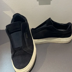 Svarta sneakers från Axel Arigato - Svarta sneakers från Axel Arigato med stilren design och vit platt sula. Skorna är tillverkade i mocka och har elastisk panel över vristen istället för snörning. Diskret logotyp i guld på sidan. Perfekt för dig som gillar minimalistisk och modern stil.