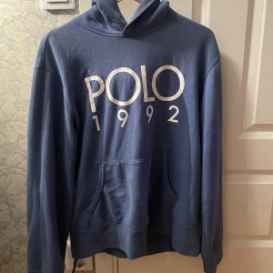 Blå hoodie från Polo Ralph Lauren - Säljer en blå hoodie från Polo Ralph Lauren med stor vit logga och årtalet 1992 tryckt på bröstet. Tröjan har huva, känguruficka och är tillverkad i mjuk bomull. Perfekt för en chill och stilren look.