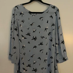 Ljusblå blus med fågelmönster från Freequent  - Supersöt ljusblå blus med svarta och vita fåglar som mönster. Blusen har 3/4  ärmar med lätt puff och knappdetalj bak i nacken. Den är rak i modellen och har en flowy känsla, perfekt för dig som gillar unika prints och en chill vibe.
