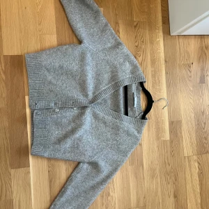 Grå kofta från Zara i ullmix - Mysig grå kofta från Zara i mjuk ullmix. Koftan har v-ringning, ribbade muddar och knäppning framtill med genomskinliga knappar. Perfekt för lager-på-lager och chill dagar.