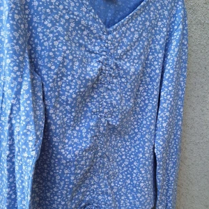 Blommig blå långärmad topp Lindex - Supersöt blå långärmad topp från Lindex med små vita blommor över hela tyget. V-ringad med rynkade detaljer framtill som ger en snygg form. Perfekt för dig som gillar en feminin och fräsch stil.