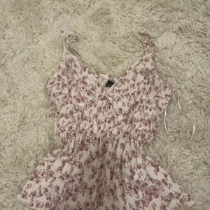 Blommig linnetopp från Gina Tricot - Supersöt linnetopp från Gina Tricot i vit med små rosa/lila blommor. Toppen har smockad midja, v-ringning och tunna axelband med knyt. Perfekt för varma dagar och ger en romantisk vibe till din outfit. Priset går att diskuteras om och skicket är bra! Är en S men passar mig superbra som en XS!💗