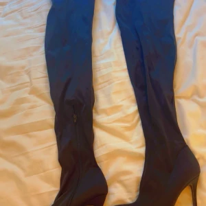 Svarta overknee stövlar med klack - Snygga svarta overknee stövlar med hög klack och dragkedja på insidan. Stövlarna har en spetsig tå och är tillverkade i ett stretchigt syntetmaterial som sitter tight längs benet. Perfekta för att lyfta vilken outfit som helst.