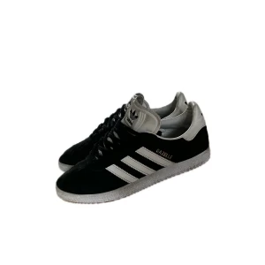 Adidas Gazelle svarta sneakers mocka - Svarta Adidas Gazelle sneakers i mocka med vita detaljer och klassiska tre ränder på sidorna. Guldtexten 'GAZELLE' på sidan och vit sula med mönster undertill. Skorna har snörning och vit plös med Adidas-logga. Perfekta för en stilren och sportig look.