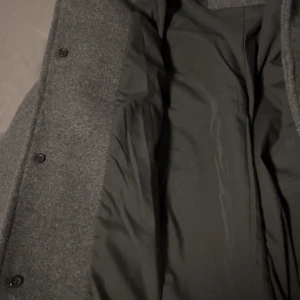 Grå ullrock med knappar - Stilren grå rock i ullmaterial med klassisk krage och stora fickor framtill. Jackan har knäppning med svarta knappar och helsvart foder på insidan. Perfekt för kyliga dagar när du vill hålla stilen enkel och clean.