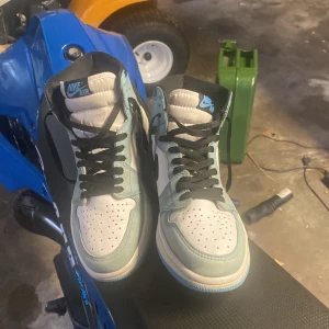 Nike Air Jordan 1 High blå/vit - Säljer ett par Nike Air Jordan 1 High med vit läderbas och ljusblå mockadetaljer. Skorna har svarta snören, svart swoosh och klassisk Air Jordan-logga på sidan. Perfekta för dig som vill sticka ut med en ikonisk sneaker i snygga färger.