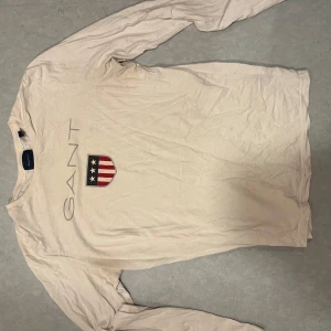 Beige/vit  långärmad tröja från GANT - Säljer en beige långärmad tröja från GANT med tryckt logga och sköld med stjärnor och ränder på bröstet. Tröjan är i mjuk bomull och har en klassisk passform som funkar till många olika stilar.