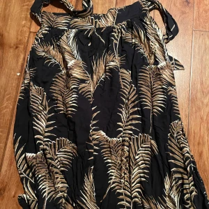 Svart långkjol med palmbladsmönster - Snygg svart långkjol med tropiskt palmbladsmönster i beige och bruna toner. Kjolen har ett snörningsband och ett luftigt, flowy fall som passar perfekt till varma dagar. Materialet känns lätt och bekvämt mot huden.