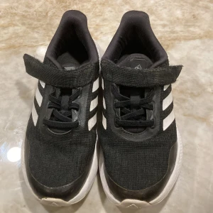 Svarta Adidas Bounce sneakers strl 31 - Svarta Adidas Bounce sneakers med vita detaljer och klassiska tre ränder på sidorna. Skorna har snörning och kardborreband för enkel påtagning. Ovandelen är i textil och syntet, sulan är vit och ger bra dämpning. Perfekta för dig som gillar sportig stil.