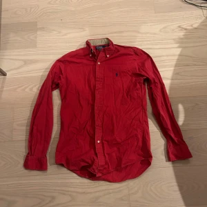 Röd skjorta från Polo Ralph Lauren - Klassisk röd skjorta från Polo Ralph Lauren med button-down krage och blå broderad logga på bröstet. Skjortan har långa ärmar, knappar framtill och är tillverkad i mjuk bomull. Perfekt för en stilren och avslappnad look.