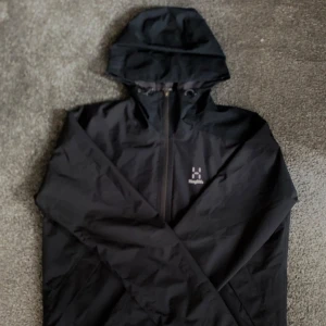 Svart vindjacka från Haglöfs med huva - Svart vindjacka från Haglöfs i storlek L. Jackan har hel dragkedja, justerbar huva och meshfoder på insidan för extra komfort. Perfekt för blåsiga dagar och outdoor-äventyr. Snygg minimalistisk design med diskret logga på bröstet.