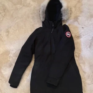 Canada goose jacka - Svart dunjacka från Canada Goose med klassisk patch på ärmen och fluffig pälskant på huvan (som är avtagbar). Jackan har mellanlång passform, dragkedja framtill och flera fickor. Perfekt för kalla vinterdagar.