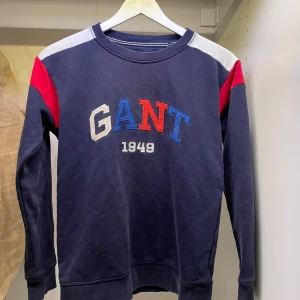 Blå Gamt tröja 146/152 - Mörkblå sweatshirt från Gant med broderad logga i flera färger framtill. Tröjan har kontrastpaneler i rött och vitt på axlarna och ärmarna vilket ger en snygg och unik design. Klassisk rund halsringning och ribbade muddar. Perfekt för en sportig och avslappnad stil. Mvh Hannes