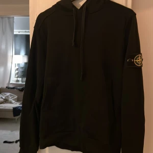 Svart hoodie från Stone Island L - Svart hoodie från Stone Island i storlek L med klassisk patch på ärmen och huva med snörning. Tröjan har en clean look och är perfekt för dig som gillar streetwear. Tillverkad i mjuk bomull för maximal komfort.