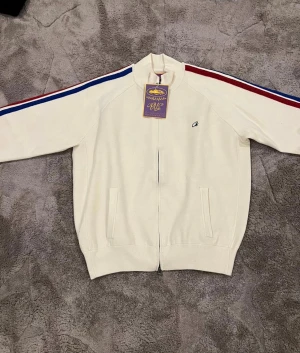 Vit track jacket  - Säljer en vit track jacket med broderad logga fram och stor graffiti-text på ryggen. Jackan har blå och röda ränder längs ärmarna, hög krage och dragkedja hela vägen. Tillverkad i mjuk bomull och har två fickor framtill.