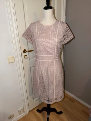 Ljusrosa spetsklänning från STOCKH LM - Supersöt ljusrosa kortklänning från STOCKH LM med vackert spetsmönster och markerad midja. Klänningen har korta ärmar, rund halsringning och dragkedja i sidan. Perfekt för dig som gillar romantisk och feminin stil.