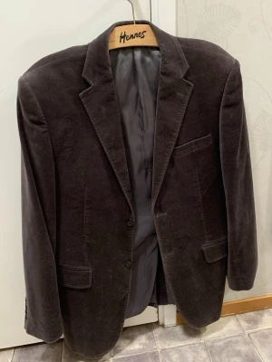 Kavaj - Säljer en grå/mörkbrun kavaj i sammets material från Batistini, storlek 52, storlek L. Klassisk modell med två knappar framtill, tre fickor och slits på baksidan. Yttertyget är 100% bomull och fodret är en mix av polyester och viskos. Perfekt för dig som vill klä upp sig och vill ha en stilren och bekväm look.