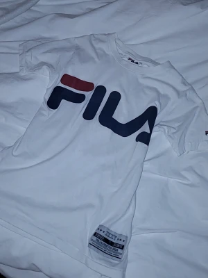 Vit FILA t-shirt med stor logga XS - Säljer en vit t-shirt från FILA i storlek XS. T-shirten har en stor FILA-logga i marinblått och rött på bröstet samt en patch nertill. Klassisk rund hals och korta ärmar. Perfekt för dig som gillar sportig och clean stil.