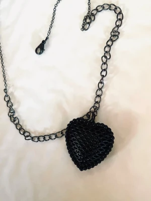 By prisförslag- Svart halsband45 cm med hjärthänge - Kedjehalsband i svart metall med ett stort hjärthänge. Hänget har ett coolt rutmönster och pärlad kant, vilket ger en edgy vibe. Perfekt accessoar för att lyfta din outfit med en unik detalj.