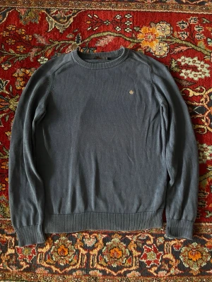 Mörkblå stickad tröja från Morris - Säljer en mörkblå stickad tröja från Morris med rund hals och diskret gul broderad logga på bröstet. Tröjan har ribbade muddar vid ärmslut och nederkant, perfekt för dig som gillar klassisk och stilren look.