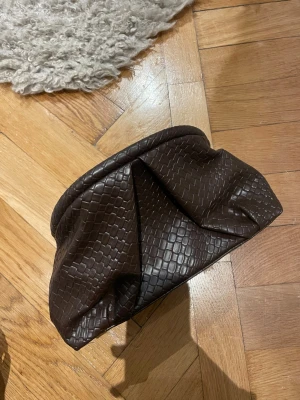 Mörkbrun kuvertväska i skinnimitation - Säljer en mörkbrun kuvertväska i skinnimitation med croco-mönster. Väskan har en mjuk, rundad form och stängs med metallspänne. Insidan är fodrad med svart tyg och har guldfärgade detaljer. Perfekt för dig som gillar stilrena accessoarer med lite edge.