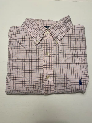 Rutig skjorta från Ralph Lauren - Säljer en klassisk rutig skjorta från Ralph Lauren i ljusrosa och blått. Skjortan har knappar framtill, button-down krage och den ikoniska broderade loggan på bröstet. Classic fit-modell i mjuk bomull, perfekt till jeans eller chinos.