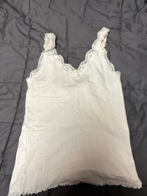 Vit spetskantad linnetopp från Cubus - Söt vit linnetopp från Cubus med tunna vertikala ränder och spetsdetaljer längs urringning och axelband. Toppen har en v-ringad form och små volangkanter nedtill. Storlek S